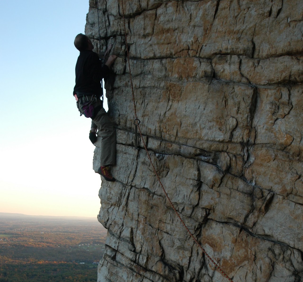 A climber ascending a sheer rock face at dusk, overlooking a wide autumn valley